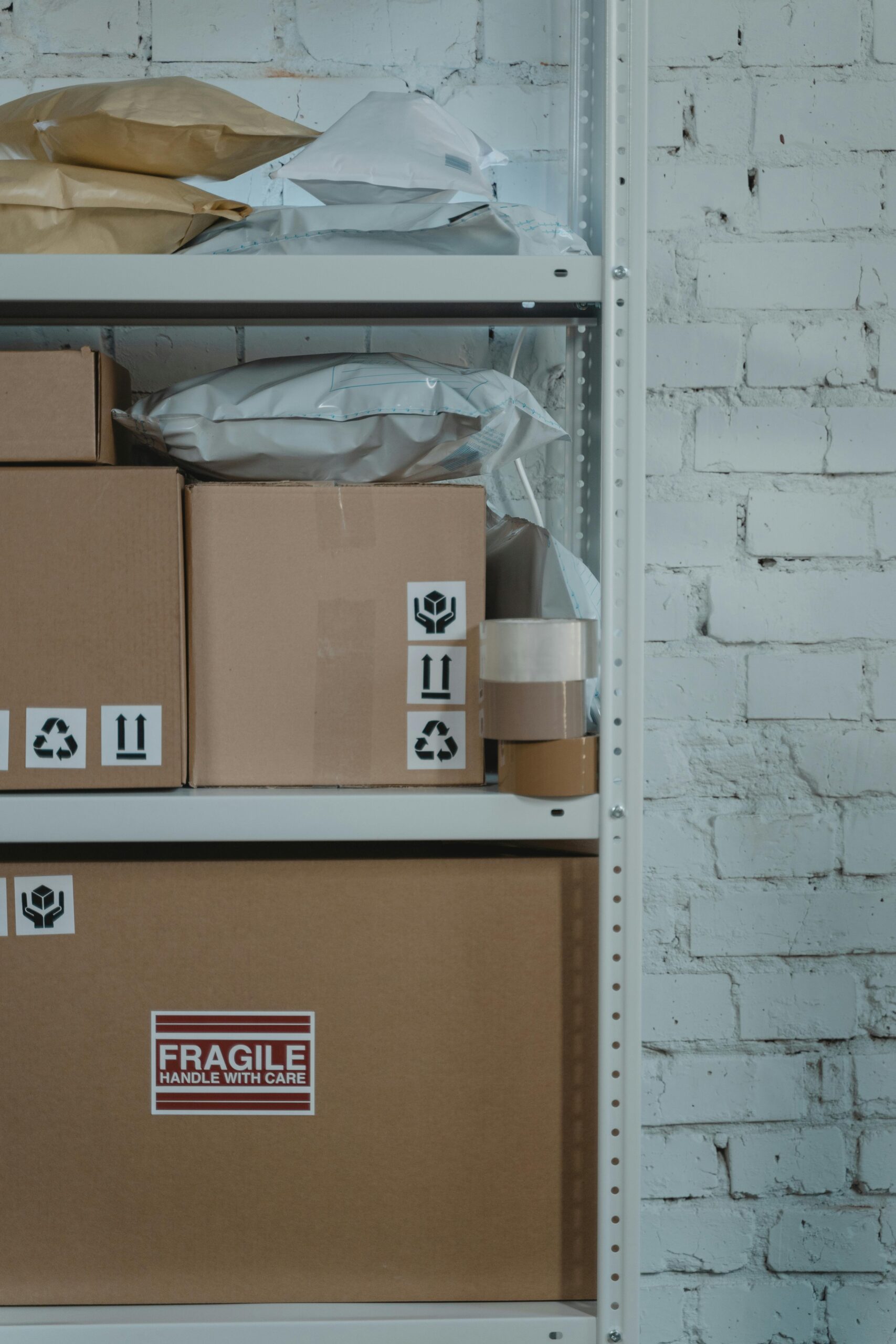 A warehouse shelf with labeled cardboard boxes and packing materials against a brick wall.